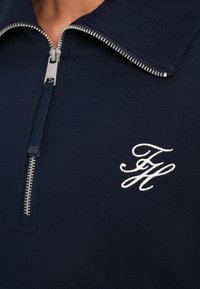 Polo bleu marine zippé avec un col montant, doté d'une fermeture éclair argentée et d'un logo brodé blanc sur le côté gauche. Texture en coton lisse.