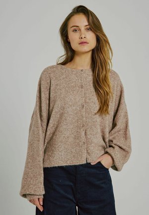 Beige neulecardigan, jossa on pyöreä kaula-aukko, nappikiinnitys ja leveät hihat. Teksturoitu kangas ja lyhyt malli yhdistettynä tummansinisiin housuihin.