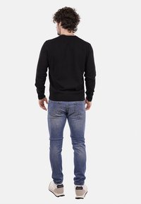 Maglione nero a maniche lunghe con polsini e orlo a costine, indossato con jeans in denim blu chiaro e sneakers di colore chiaro, mostrato da dietro.