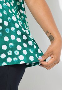 Groene top met witte en turquoise cirkelpatronen. De stof heeft een gladde textuur. Een hand trekt aan de onderrand.