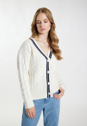 Junge Frau mit langen, welligen Haaren, die eine weiße, kabelgestrickte Strickjacke mit navyfarbenem Rand und Knöpfen trägt, kombiniert mit hellblauen Jeans.