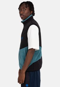 Black and teal color-blocked vest with high collar, featuring a zipper, side pockets, and blue logo detail. Worn over a white shirt.