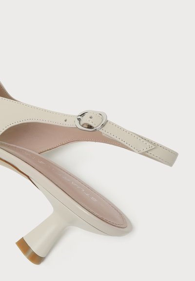 Μπεζ δερμάτινο παπούτσι slingback με καμπυλωτή μεσαίου ύψους φτέρνα και ασημένιο λουράκι με αγκράφα σε λευκό φόντο.