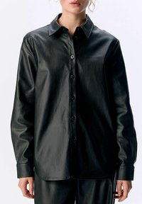 Chemise noire en simili cuir à boutons, avec un col classique, des manches longues et une texture lisse ; présente des boutons noirs sur le devant.