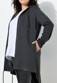 Giacca lunga nera con cappuccio, dal taglio rilassato, tasche laterali e orlo posteriore leggermente più lungo, indossata sopra una maglietta bianca semplice.