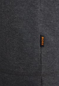 Tissu texturé gris foncé avec une couture visible ; présente une petite étiquette noire avec le mot "BOSS" en orange.