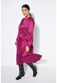Fuchsia langärmliges Kleid mit gestuftem Design, strukturierter Musterung und hohem Rundhals, kombiniert mit schwarzen kniehohen Stiefeln und Creolen.