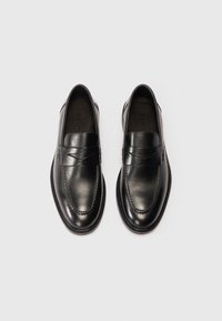Uz melna āda pulētām slip-on loafers, ar izšuvumiem, skatīts no augšas uz balta fona.