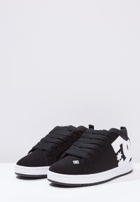 Mustat tennarit teksturoidulla päällisellä, valkoisella logolla sivulla ja valkoisilla kumipohjilla. Varustettu pehmustetulla kielekkeellä ja nauhoilla tukevan istuvuuden takaamiseksi.