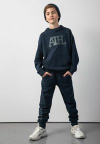 Granatowy zestaw dresowy z bluzą z kapturem, z dużym graficznym logo na górze. Połączony z białymi butami sportowymi. Miękki, teksturowany materiał.