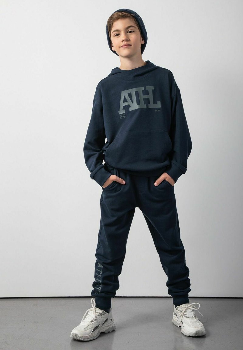 Granatowy zestaw dresowy z bluzą z kapturem, z dużym graficznym logo na górze. Połączony z białymi butami sportowymi. Miękki, teksturowany materiał.