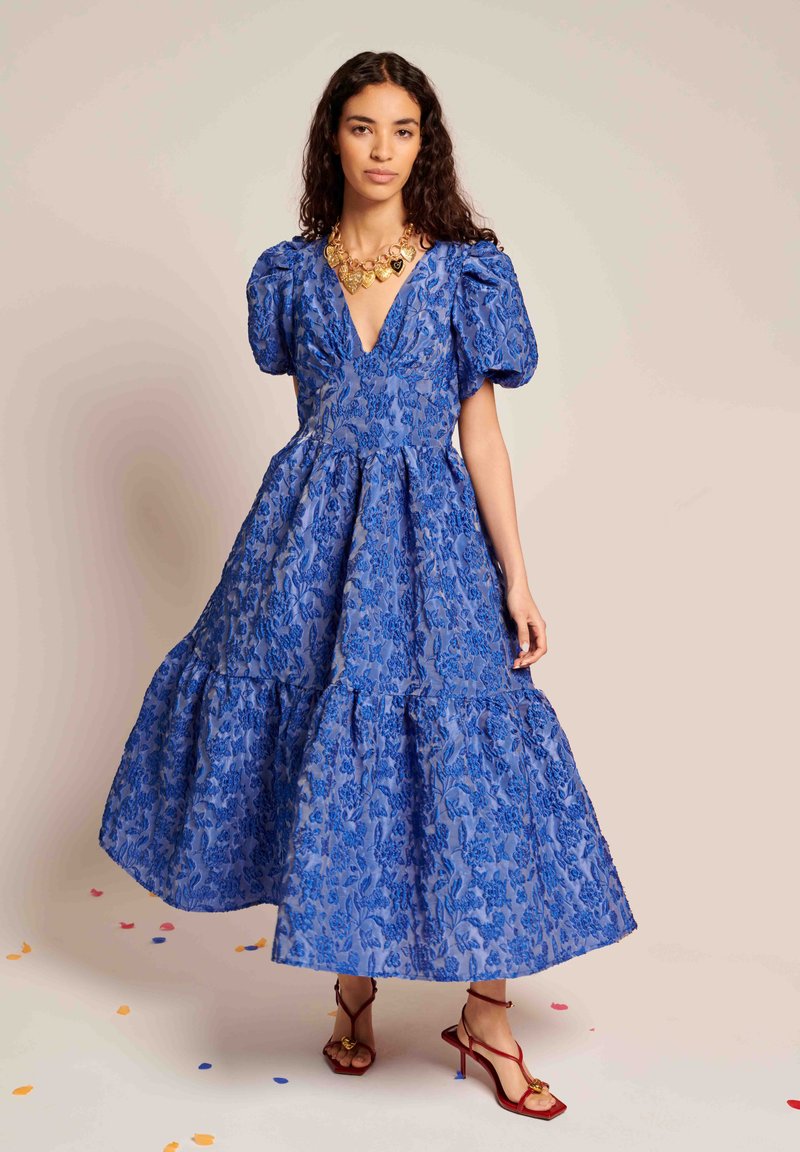 Μπλε φόρεμα με λουλουδάτο μοτίβο, φουσκωτά μανίκια, βαθύ V-λαιμόκοψη και πολλαπλά στρώματα. Συνδυασμένο με κόκκινες σαμπό με λουράκια και χρυσό εντυπωσιακό κολιέ.