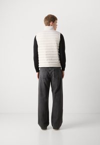 Lättvikts pufferväst i off-white med en quiltad design, hög krage och en slät textur, bärs över en svart långärmad skjorta.