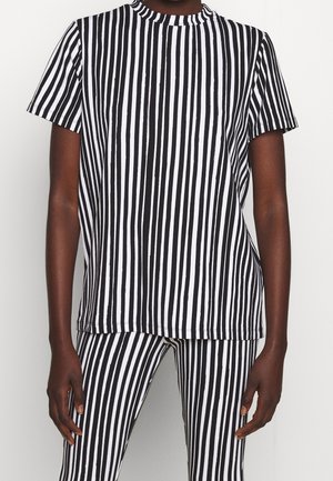 Person trägt ein kurzärmeliges schwarz-weiß gestreiftes Hemd mit vertikalen Streifen und passende vertikal gestreifte Hosen vor einem schlichten Hintergrund.