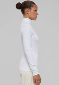 Maglione bianco a collo alto con un design aderente, texture morbida, maniche lunghe e polsini a coste, indossato con pantaloni di colore chiaro.