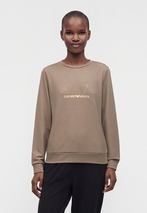 Ung person med kort hår iført en beige sweatshirt med EA7 Emporio Armani-logo i guld, stående mod en ensfarvet hvid baggrund.