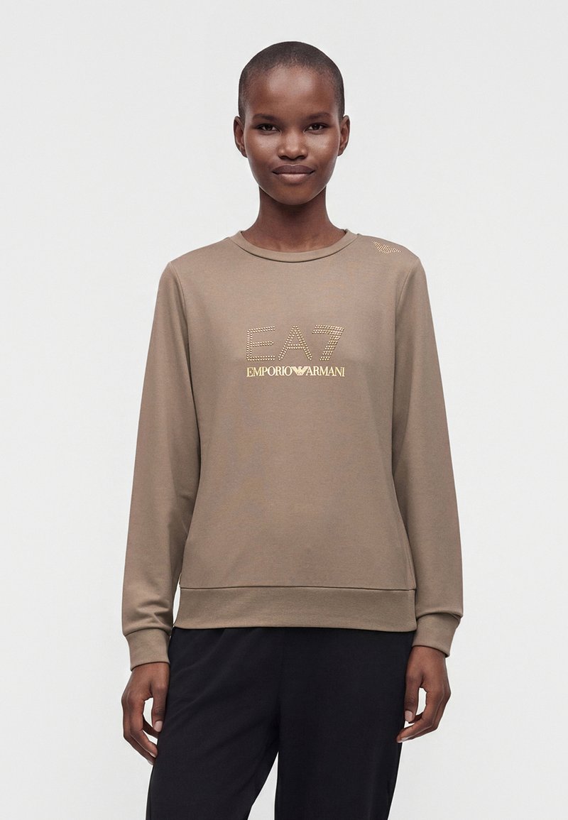 Jeune personne aux cheveux courts portant un sweat-shirt beige avec le logo EA7 Emporio Armani en or, debout devant un fond blanc uni.