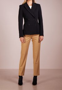 Kvinna iklädd en åtsittande svart dubbelknäppt kavaj, beige skräddarsydda byxor och svarta ankelsneakers mot en neutral bakgrund.