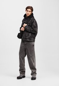 Giacca nera con motivo e cappuccio, jeans grigi larghi e stivali neri. Mostra un design intricato e texture contrastanti.