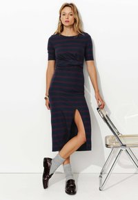 Robe rayée marine à manches courtes, avec fente sur le côté et design ajusté. Associée à des chaussettes grises et des chaussures bordeaux brillantes. Fond clair.
