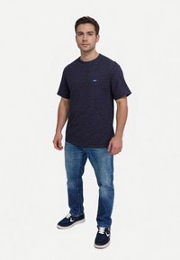 Short-sleeved navy shirt with white and red polka dots, featuring a small logo patch on the chest, paired with blue jeans and dark sneakers.