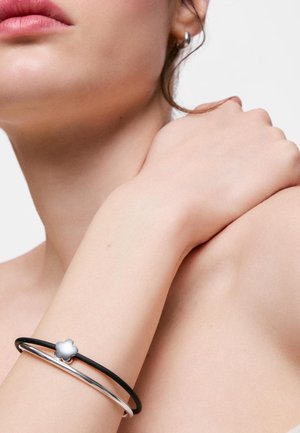 Primer plano del cuello, la barbilla y el hombro de una persona que lleva una pulsera plateada y una pulsera negra con un adorno en forma de flor plateada.