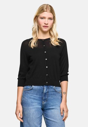 Woman with long blonde hair wears a black buttoned cardigan and blue jeans, standing with neutral expression on plain background.
