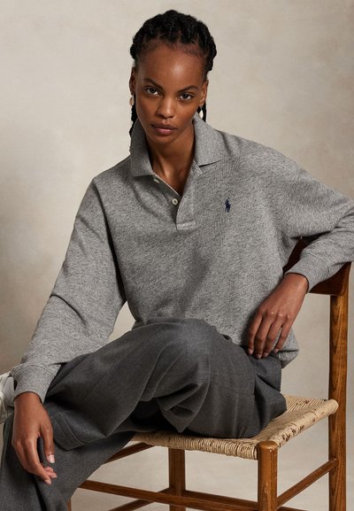 Woman with braided hair wearing a gray long-sleeve polo and dark gray pants, seated on a wooden chair with woven seat.