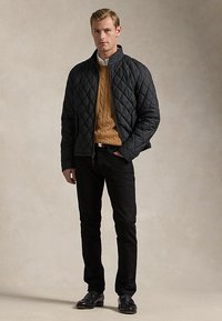 Mann iført en svart vattert jakke over en brun kabelstrikket genser, svarte bukser og svarte loafers, stående med en hånd i lommen.