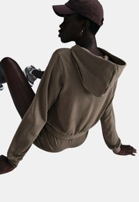 Felpa sportiva marrone con cappuccio regolabile e vita elastica, abbinata a un pantalone coordinato. La texture appare liscia e morbida.