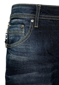 Jeans de mezclilla azul oscuro con costuras amarillas, un botón plateado y un pequeño detalle en el bolsillo trasero. Textura suave con un desvanecimiento sutil.