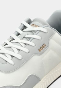 Sneaker bianca e grigia chiara con un tessuto testurizzato, lacci e branding sottile; presenta una punta arrotondata e una suola in gomma con motivo.