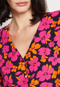 Kwiatowa sukienka z ciemnonawym tłem, zdobiona dużymi, żywymi różowymi i pomarańczowymi kwiatami. Dekolt w kształcie litery V i detale z guzikami. Miękka tekstura materiału.