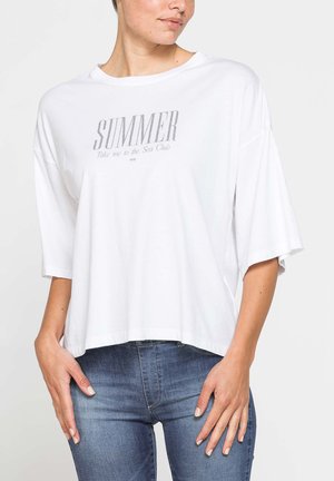 Donna che indossa una T-shirt bianca larga con il testo "SUMMER Take me to the Sea Club" e jeans blu aderenti, in piedi con le mani vicino ai fianchi.