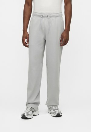 Homme portant un pantalon de survêtement gris clair avec cordon de serrage et des baskets athlétiques gris argenté, debout devant un fond blanc uni.