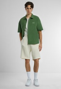 Camicetta verde a maniche corte con taschino, abbinata a pantaloni corti testurizzati color crema. Sneakers bianche completano l'outfit.