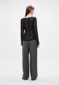 Μαύρη διαφανής μπλούζα με λεπτομερείς ασημένιες ανθισμένες παραστάσεις, συνδυασμένη με γκρίζες παντελόνες φαρδιών ποδιών με ρίγες. Λείος υφασμάτινος χαρακτήρας και εφαρμοστό σχέδιο.