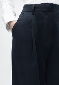 Παντελόνια σε σκούρο μπλε με εφαρμογή κομμένη στα μέτρα που διαθέτουν απαλό ύφασμα, φαρδιά ζώνη μέσης, πλαϊνές τσέπες και άνετη γραμμή, αναδεικνύοντας διακριτικά τις πτυχώσεις του υφάσματος.