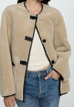 Beige zachte jas met zwarte bies en knopen over een witte cropped top, gecombineerd met high-waisted blauwe jeans, hand rustend in de zak.