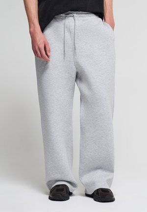 Persona che indossa pantaloni della tuta larghi grigio chiaro con coulisse in vita e sneakers nere, in piedi con una mano in tasca.
