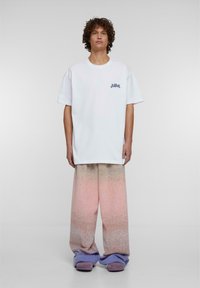 Balta per didelį t-shirt su tamsiai mėlynu logo, derinama su pastelinių spalvų gradientiniais plačiais kelnais. Minkšta tekstūra, žemas juosmuo ir purpuriniais akcentais puošti bateliai.