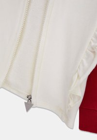 Weiße Jacke mit Reißverschluss und strukturiertem Innenfutter, ausgestattet mit einem silbernen Reißverschluss und Rüschenakzenten auf einer Seite, kombiniert mit einem roten, gerippten Bündchen.