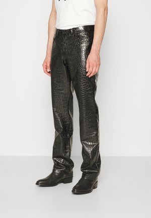 Mężczyzna ubrany w błyszczące czarne spodnie ze skóry z tłoczeniem imitującym krokodyla, czarne buty i biały bezrękawnik, stojący na jednolitym tle.