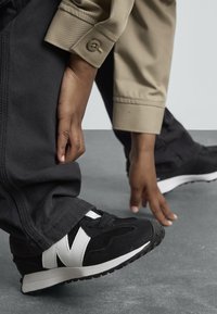 Baskets de sport noir et blanc en mesh et daim, avec un logo blanc prominent, sur une surface en béton avec un pantalon noir.