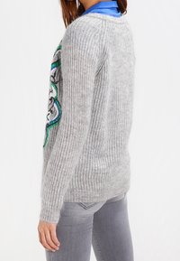 Pull gris tricoté avec texture côtelée, présentant un motif brodé coloré en bleu, vert et noir sur la manche gauche.