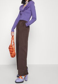 Femme portant un cardigan à col duveteux violet, pantalon marron taille haute, chaussettes florales colorées, talons plateformes lavande, tenant un sac à main à carreaux orange.