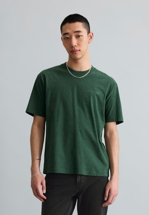 Jonge man met kort haar draagt een groen T-shirt met korte mouwen, zwarte broek, zilveren ketting en armband, staande tegen een effen grijze achtergrond.