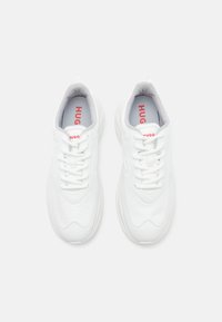 White sneakers with a textured upper, lace closure, padded collar, and red "HUGO" branding on the tongue and insole. Rounded rubber sole.