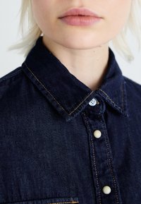 Denimová košeľa v tmavomodrej farbe s kontrastným oranžovým prešívaním, klasickým golierom a zapínaním na gombíky s bielymi detailmi.
