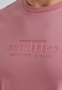 Růžové bavlněné tričko s vyraženým nápisem "NEOMEZENÁ SVOBODA PM LEGEND AMERICAN CLASSIC" na přední straně, s designem klasického kulatého výstřihu.